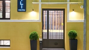 Un bâtiment jaune avec une porte noire et deux plantes en pot devant l’entrée.