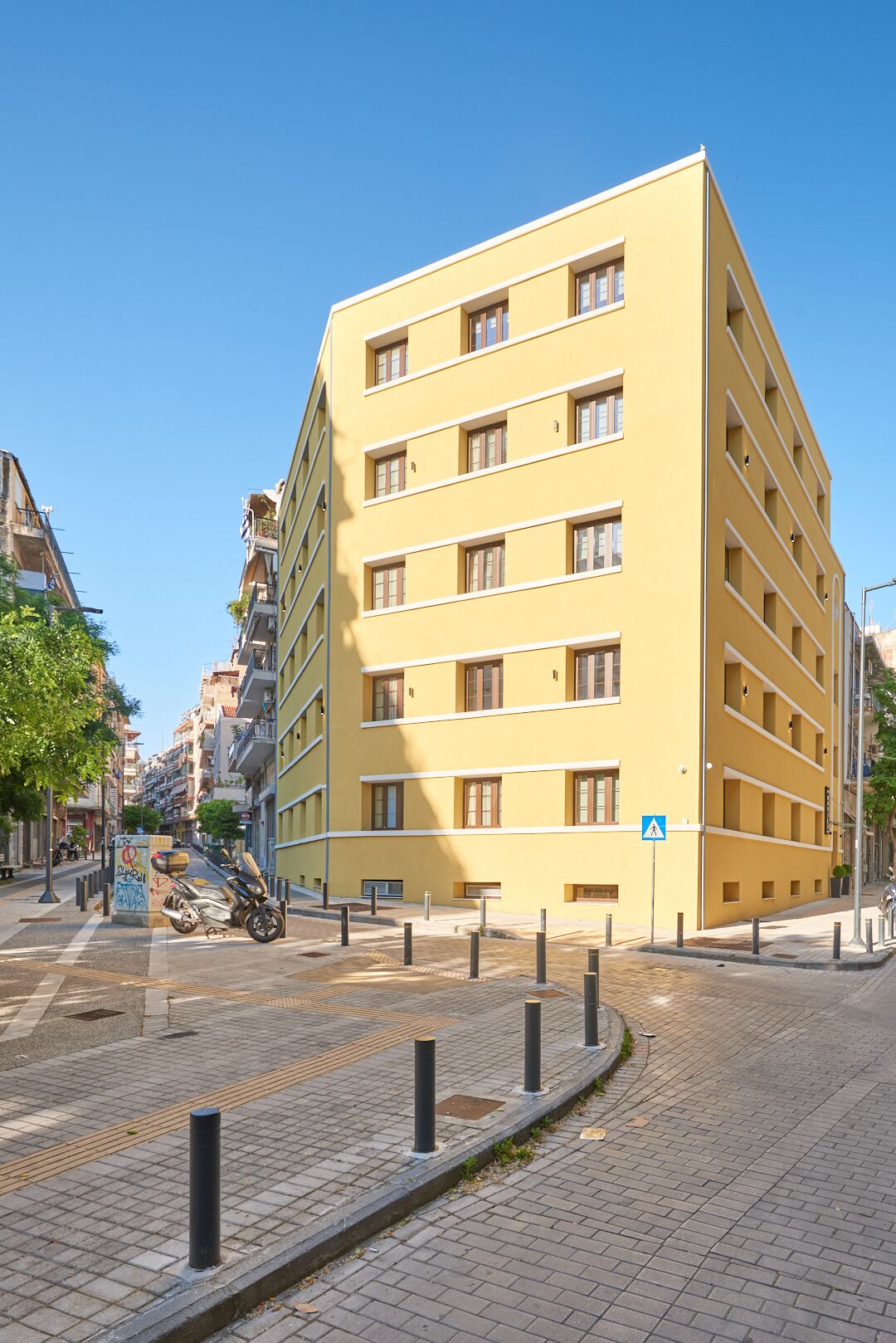 A yellow building with multiple windows and balconies, located on a paved street with bollards and a pedestrian crossing.