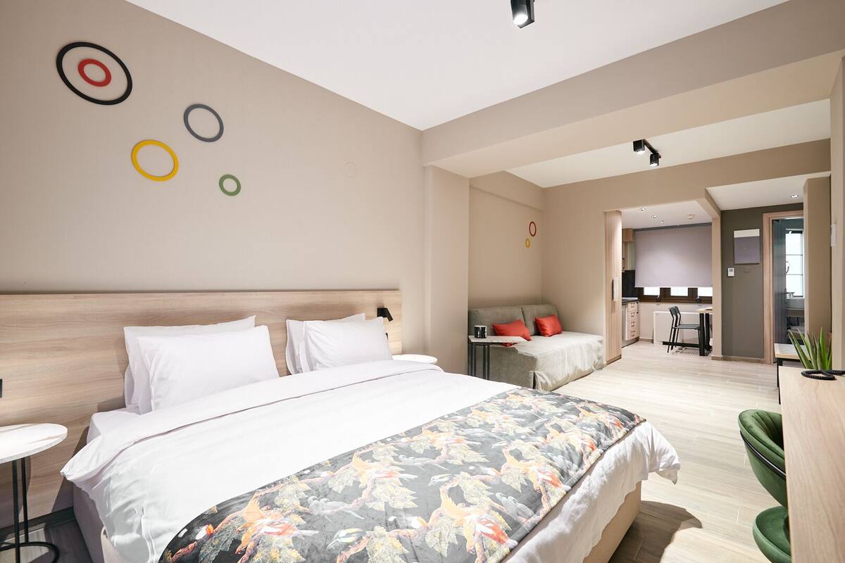 A modern hotel room with a large bed, a small table, a sofa, and a kitchenette in the background.