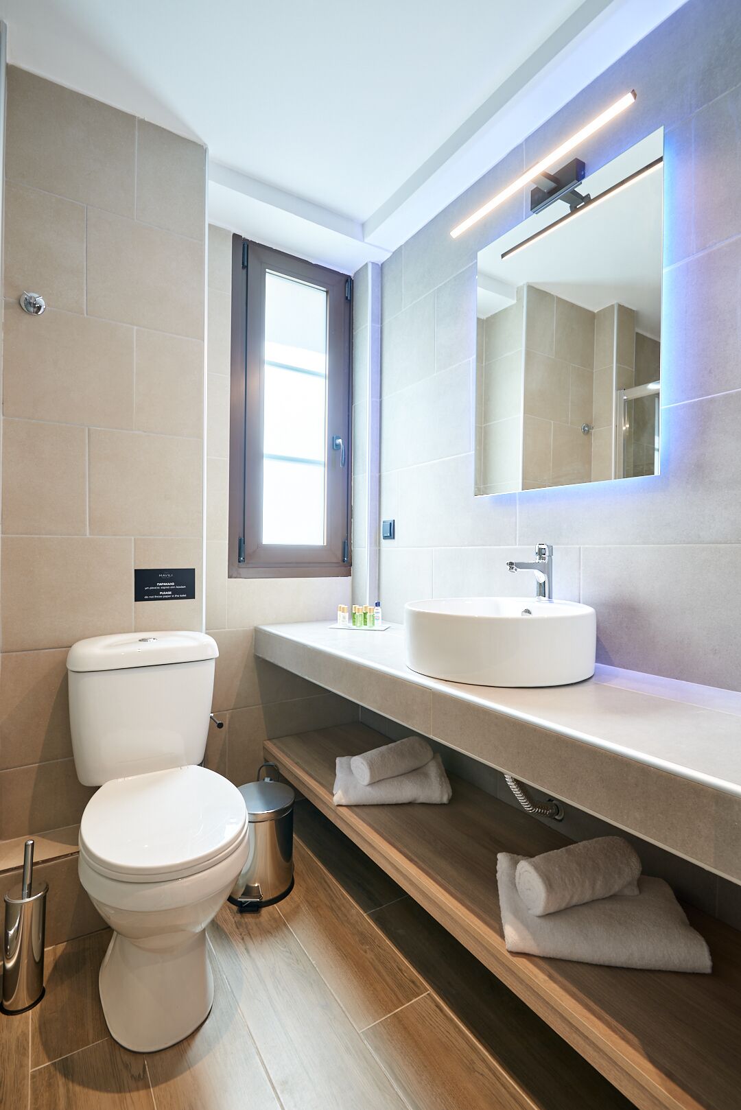A modern bathroom with a toilet, sink, and mirror. There is a window providing natural light and a shelf with towels.