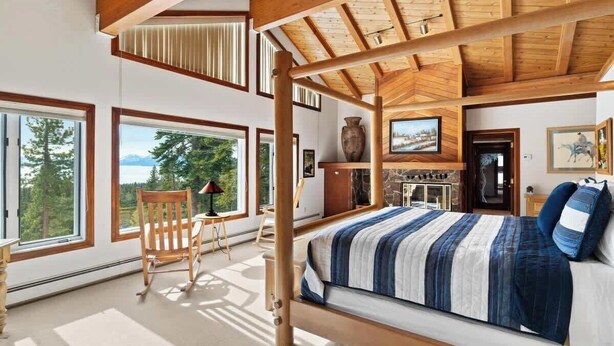 Ein Schlafzimmer mit einem großen Bett, Holzbalken an der Decke, Blick ins Freie und einem Kamin aus Stein.