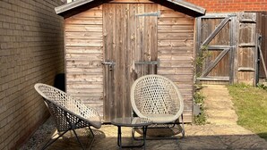 Een houten tuinhuis met een metalen deur, twee rieten stoelen en een kleine tafel in een achtertuin.