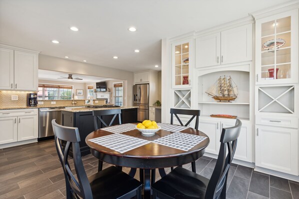 A modern kitchen with white cabinets, a central island with a bowl of lemons, and a dining area with four chairs. - Updated 4 BD, private pool, screened porch, walk to beach, waterway view! (Isle of Palms)