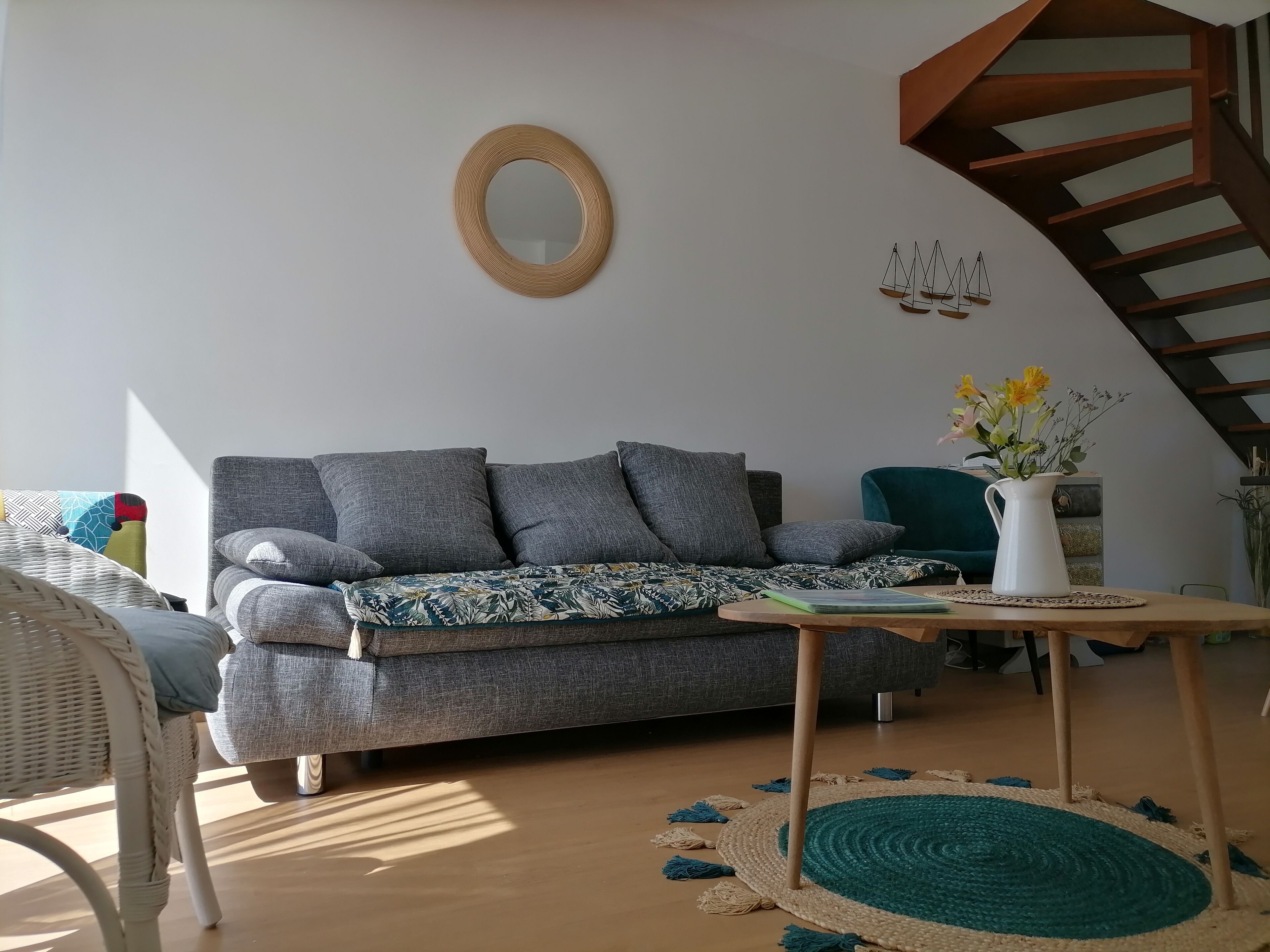 Een woonkamer met een grijze bank, een rieten fauteuil, een houten salontafel met een bloevas, en een trap met houten leuningen.