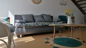 A living room with a grey sofa, a wicker chair, a wooden coffee table with a vase of flowers, and a staircase with wooden railings. - Maison à la Mer, "Le Petit Léjon", Idéale Familles ou Amis (Plérin)