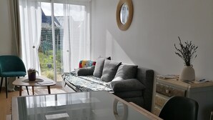 A modern living room with a glass-top dining table, a grey sofa, and a view of a garden through a sliding glass door. - Maison à la Mer, "Le Petit Léjon", Idéale Familles ou Amis (Plérin)