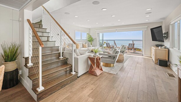 A spacious living area with a wooden staircase, a large window overlooking the sea, and a flat-screen TV mounted on the wall.