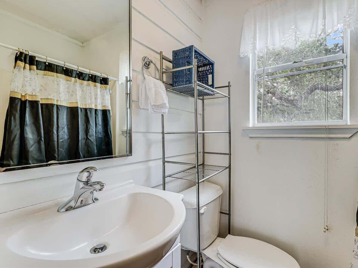 A bathroom with a white sink, a mirror, a toilet, a window with a view of trees, and a metal shelving unit with a blue basket.