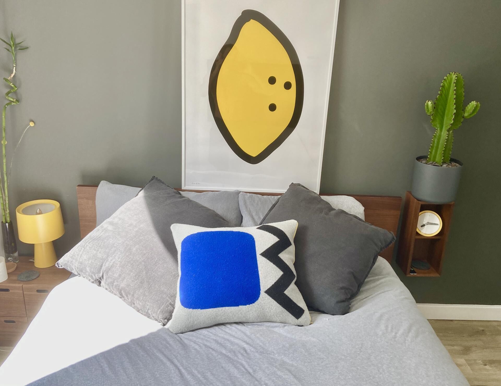 Una camera da letto con un letto, cuscini, un quadro raffigurante un avocado, una pianta di cactus, una lampada e una sveglia.