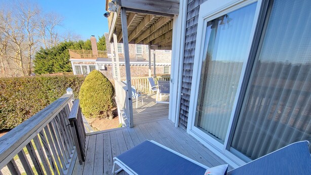 Une terrasse en bois avec un fauteuil de relaxation et une petite table, donnant sur un jardin avec une haie et une maison en arrière-plan.