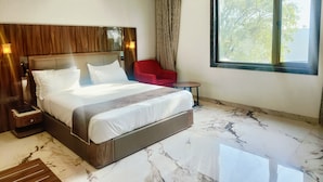 Une chambre d’hôtel avec un grand lit, un fauteuil rouge, une petite table et une fenêtre donnant sur des arbres.