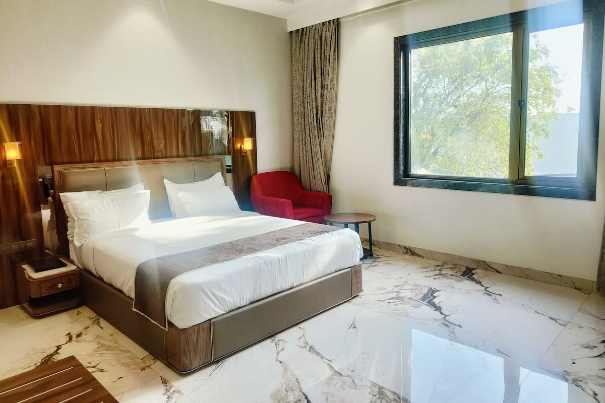 Une chambre d’hôtel avec un grand lit, un fauteuil rouge, une petite table et une fenêtre donnant sur des arbres.