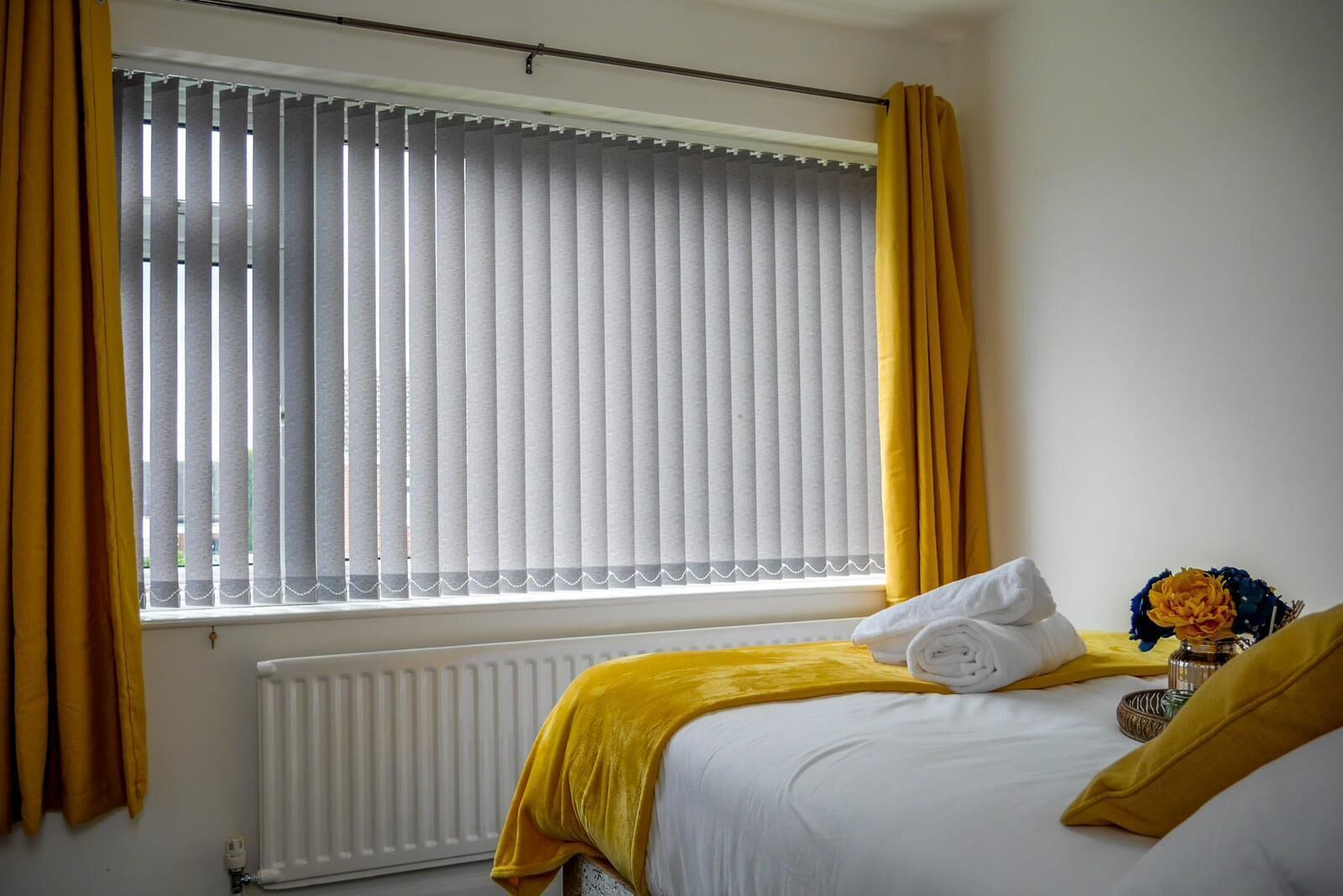 A hotel room with a bed, a radiator, a window with blinds, and a vase with flowers.