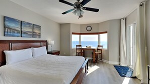 Una habitación de hotel con una cama grande, un ventilador de techo, una mesa de comedor con sillas, vista al océano y un reloj en la pared.