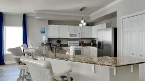 Una cocina con armarios blancos, encimera de granito, refrigerador de acero inoxidable y un taburete de bar con cojín estampado.