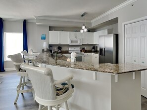 Eine Küche mit weißen Schränken, einer Arbeitsplatte aus Granit, einem Edelstahlkühlschrank und einem Barhocker mit gemustertem Sitzkissen.