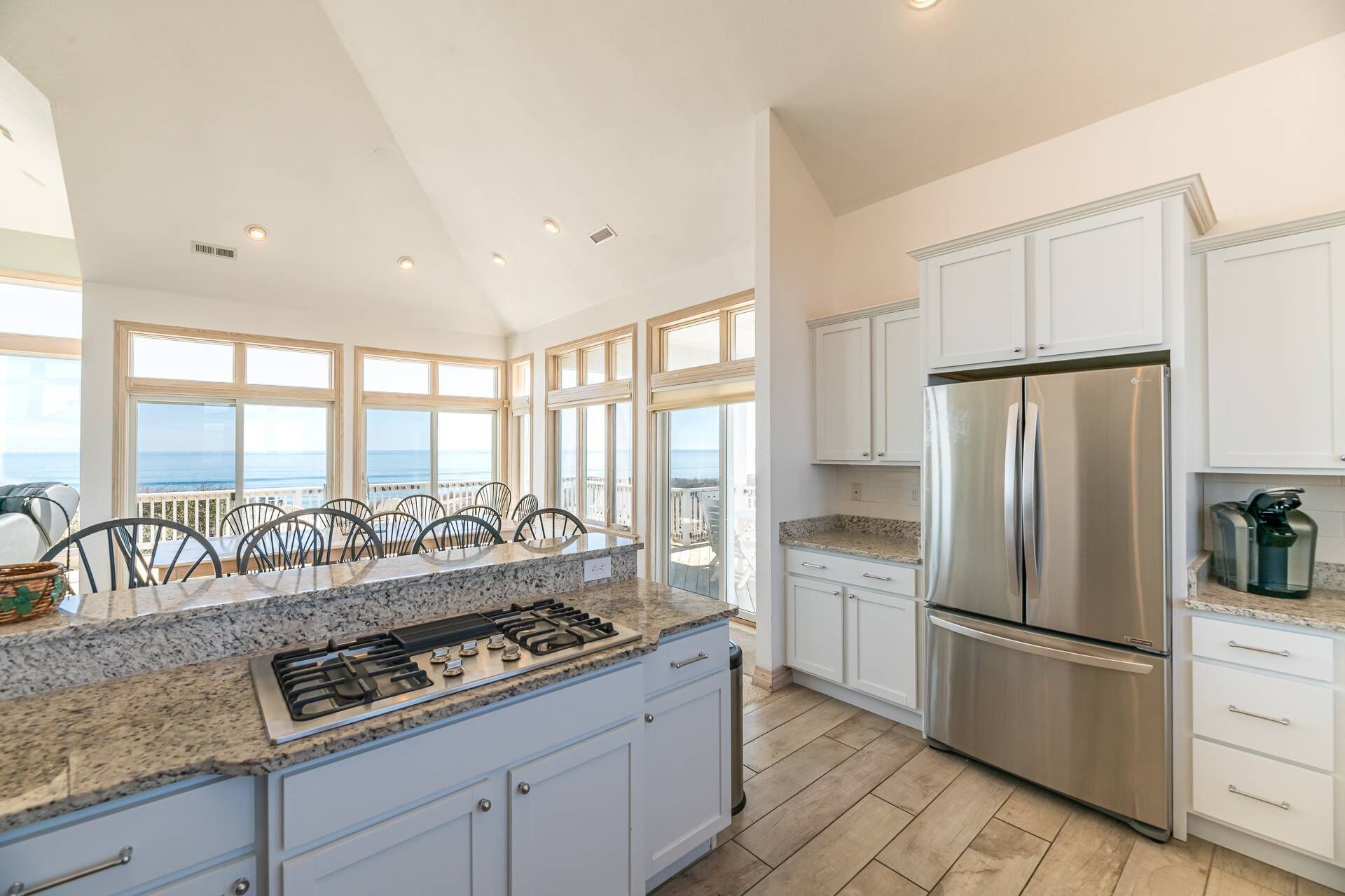 A modern kitchen with a stainless steel refrigerator, granite countertops, and a view of the ocean through large windows.