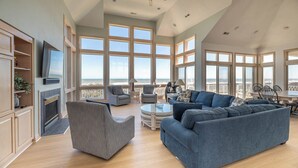 Een ruime woonkamer met een grote bank, fauteuils en een televisie. De kamer heeft grote ramen met uitzicht op het strand.