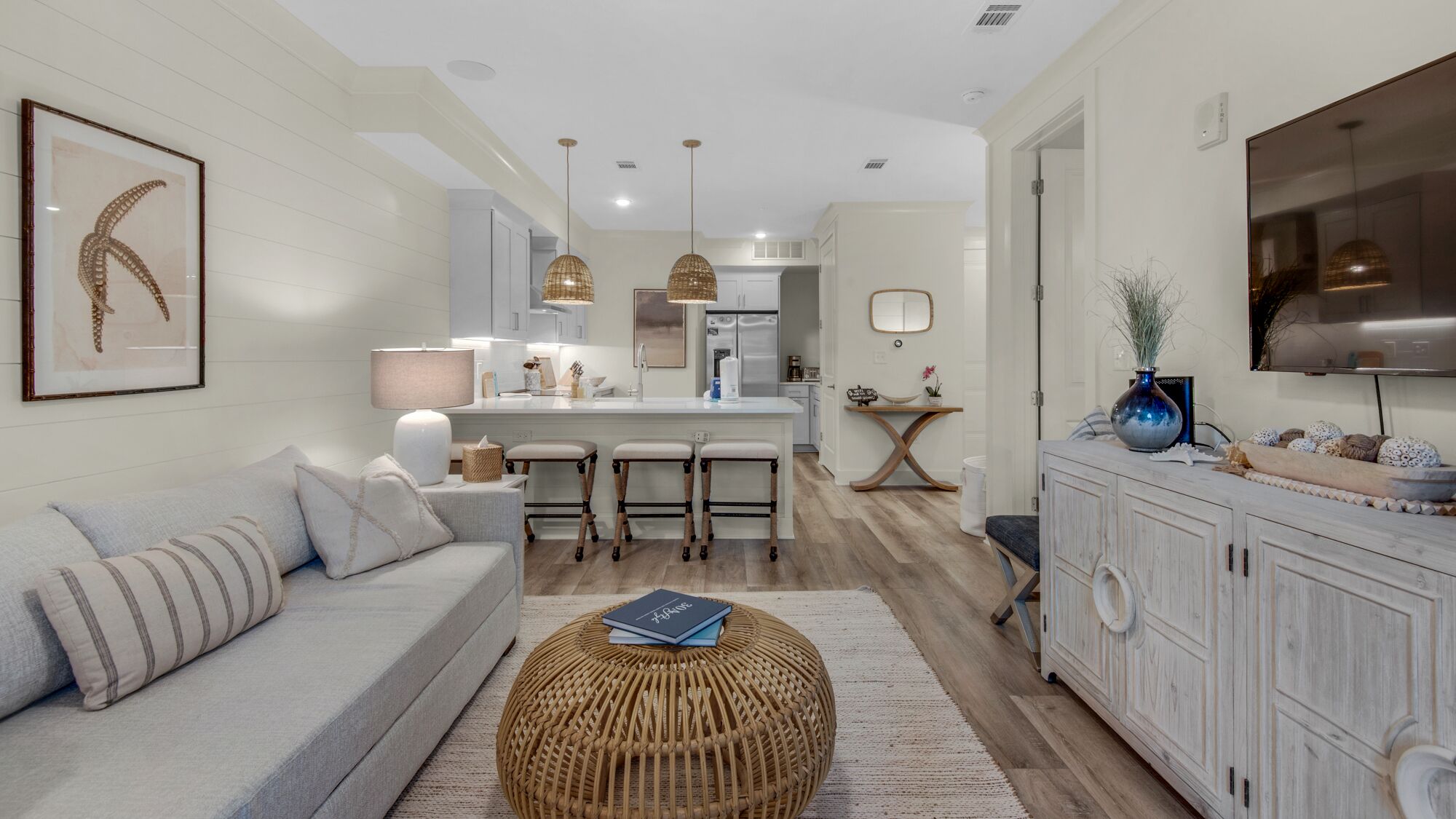 A modern living room with a sofa, a round wicker ottoman, and a dining area with a kitchen in the background.