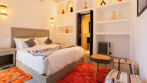 A hotel room with a bed, bedside tables, a nightstand, a chair, a small table, a radiator, and decorative items on shelves.