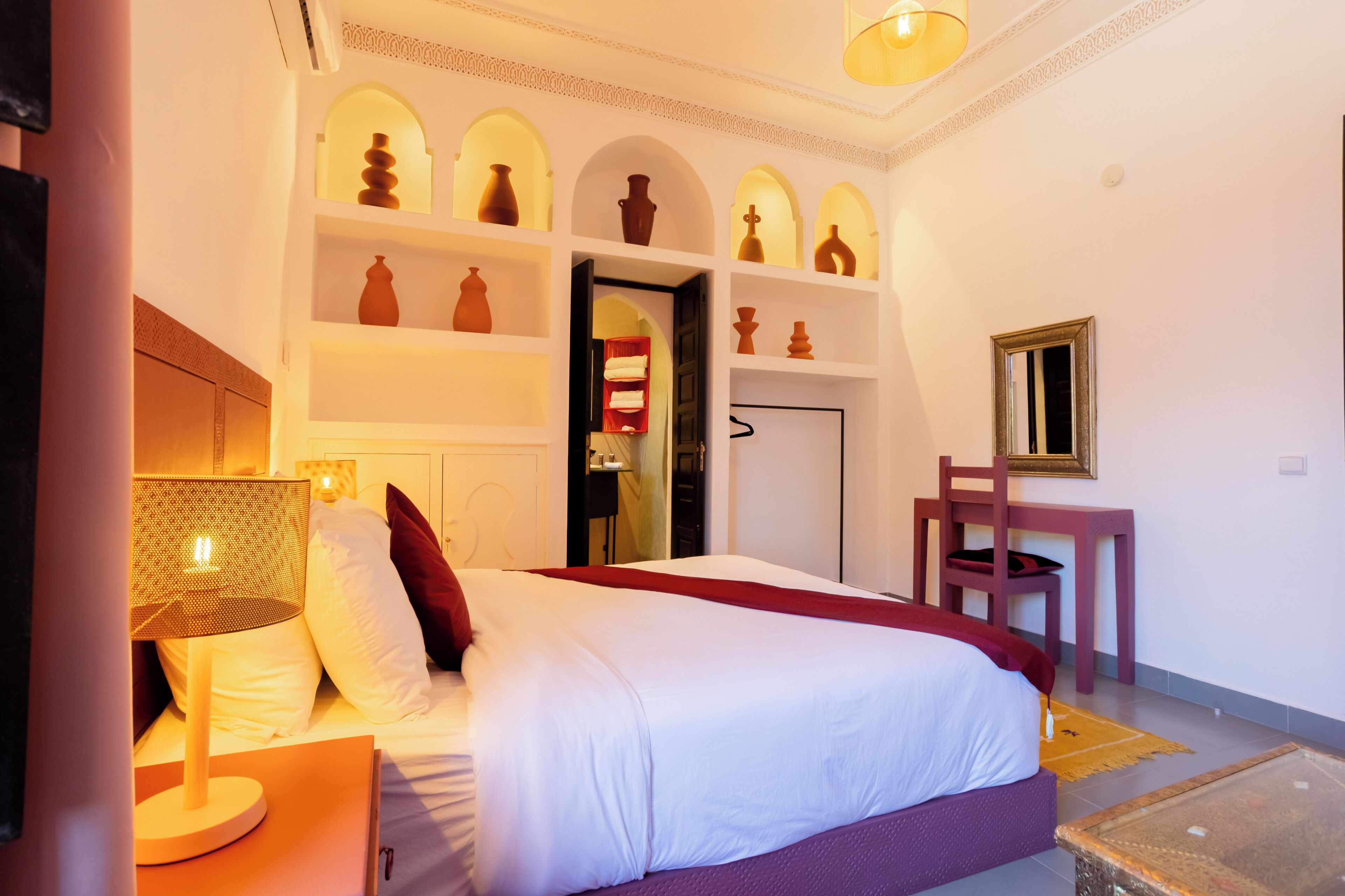 Una habitación de hotel con cama, mesita de noche, lámpara y estantes con objetos decorativos.