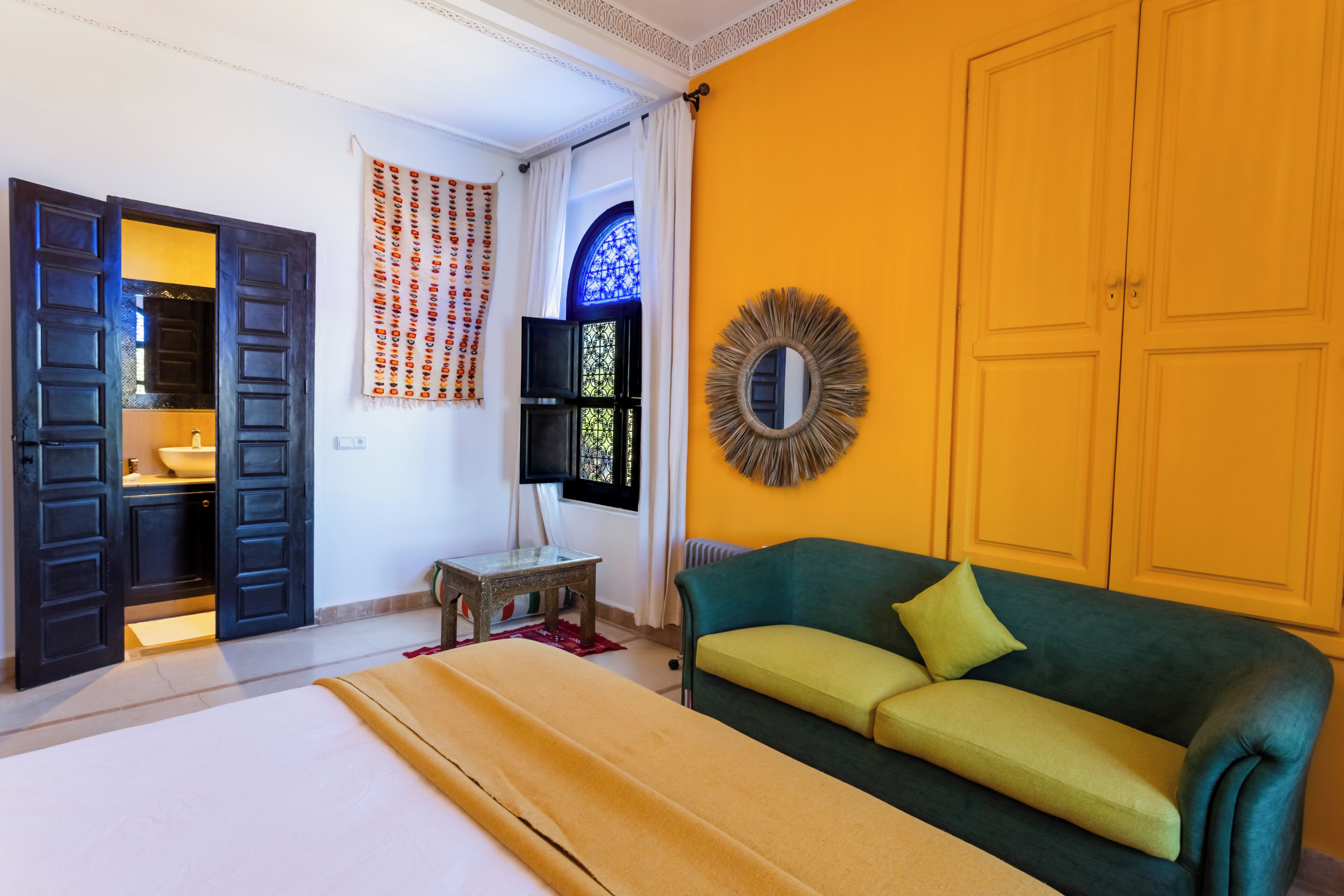 Una habitación con cama, sofá verde, espejo, ventana con cortinas y una puerta.
