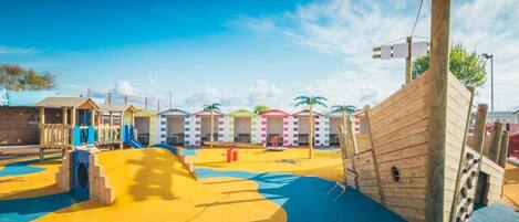A playground with colorful play structures and a slide, surrounded by small beach huts.