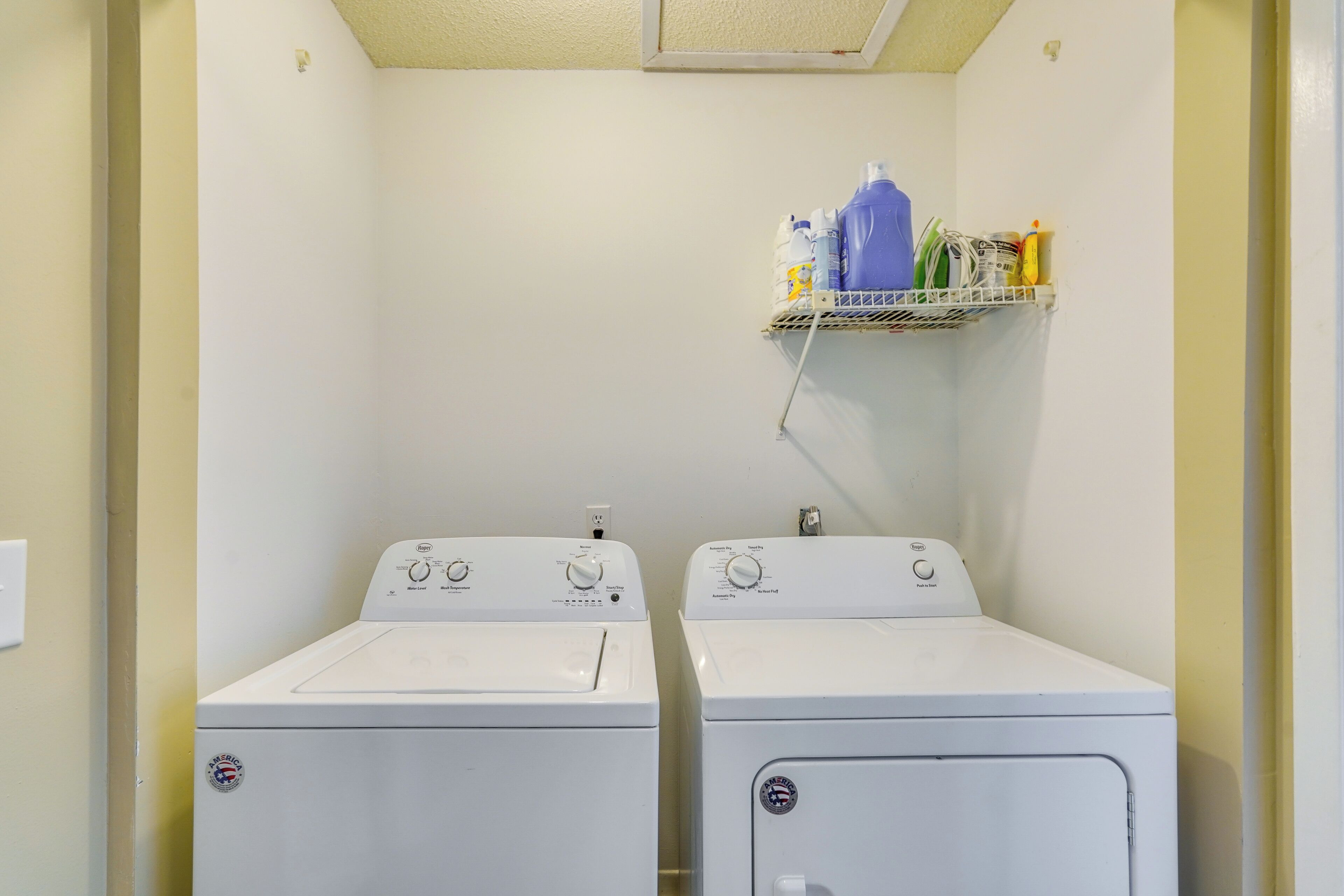 Un cuarto de lavandería con dos lavadoras de carga frontal blancas y un estante superior con productos de limpieza.