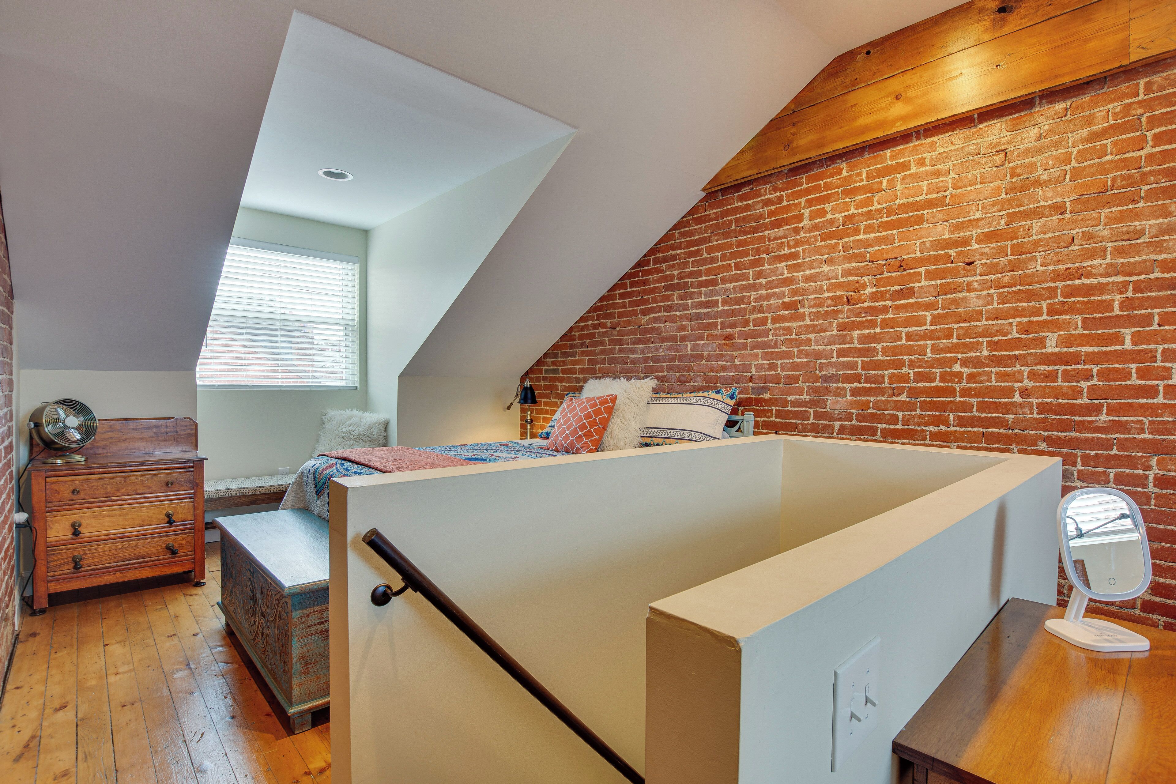 Une chambre à coucher aménagée dans un espace mansardé, avec un plafond en pente, un mur en briques apparentes, une commode en bois et un petit bureau doté d’un miroir.