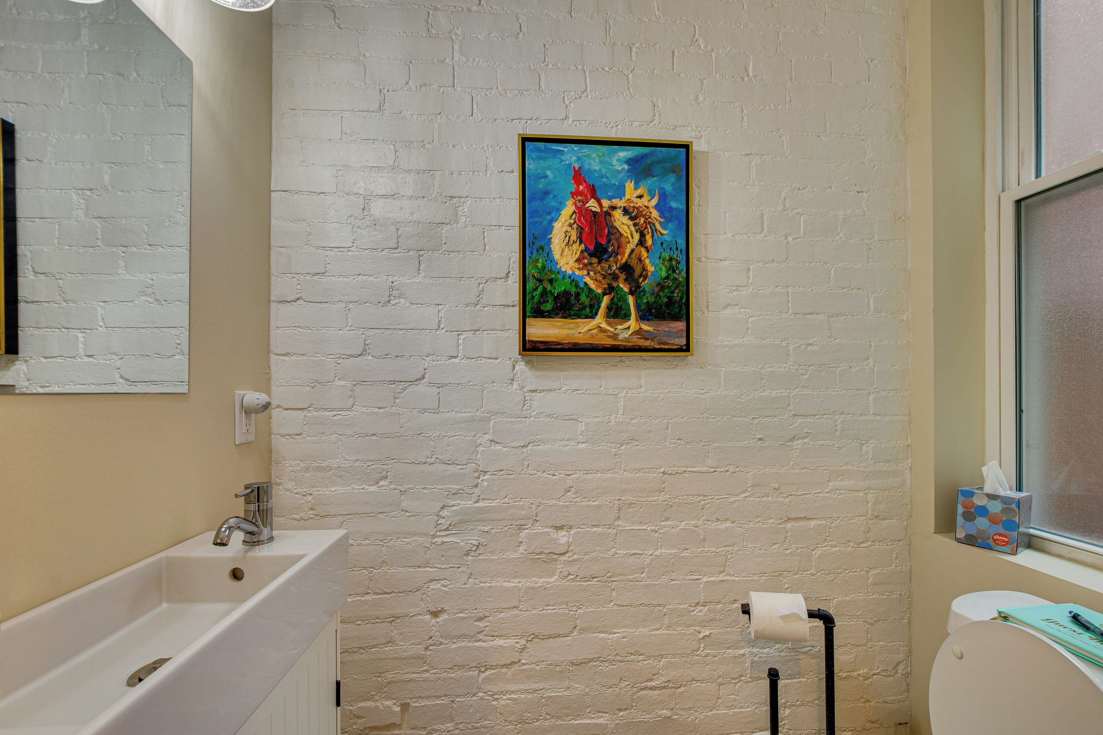 A bathroom with a white brick wall, a sink, a toilet, and a painting of a rooster.