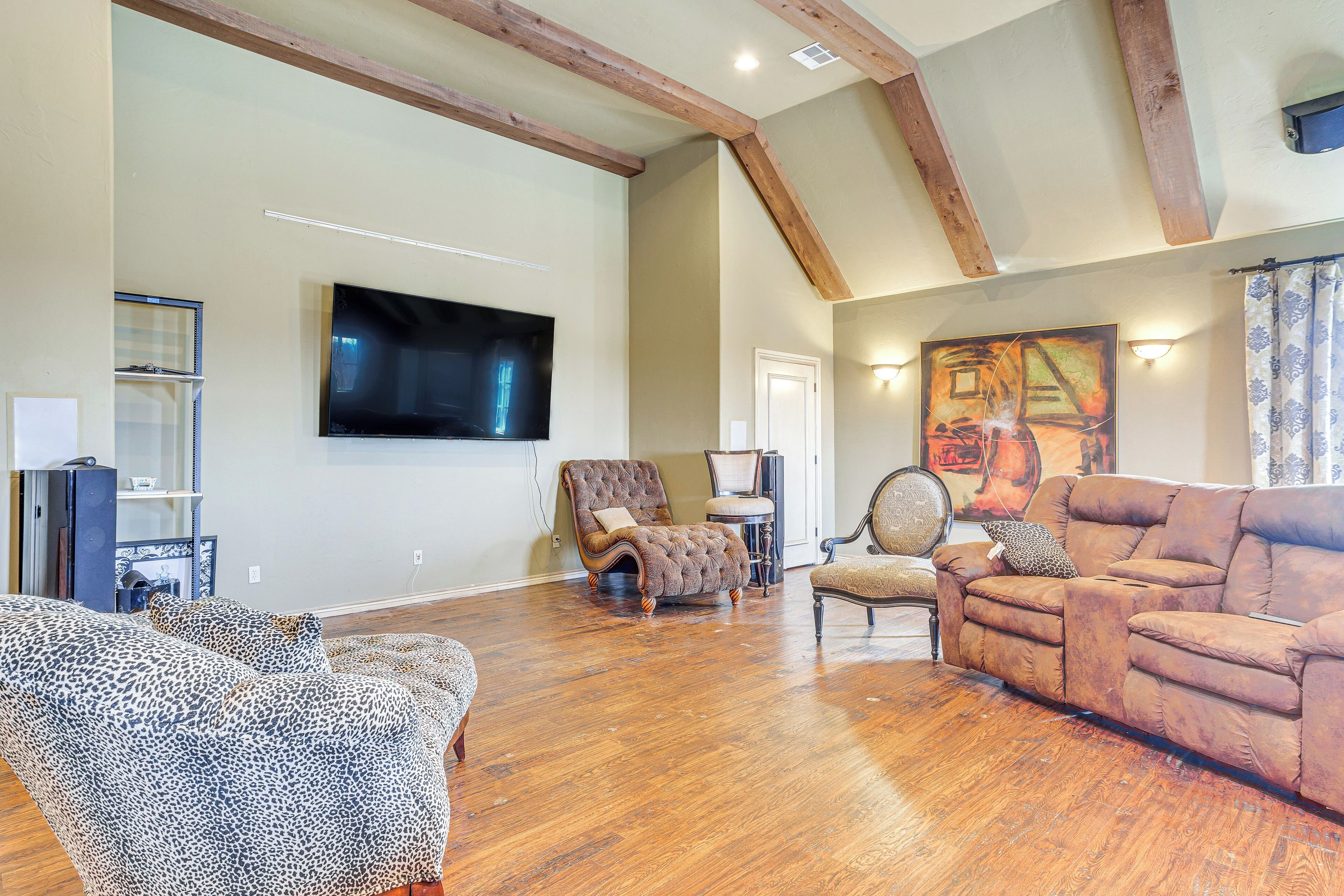 A spacious living room with hardwood flooring, a large flat-screen TV mounted on the wall, and a cozy seating area with a brown sofa and patterned armchairs.