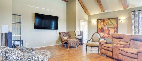 A spacious living room with hardwood flooring, a large flat-screen TV mounted on the wall, and a cozy seating area with a brown sofa and patterned armchairs.