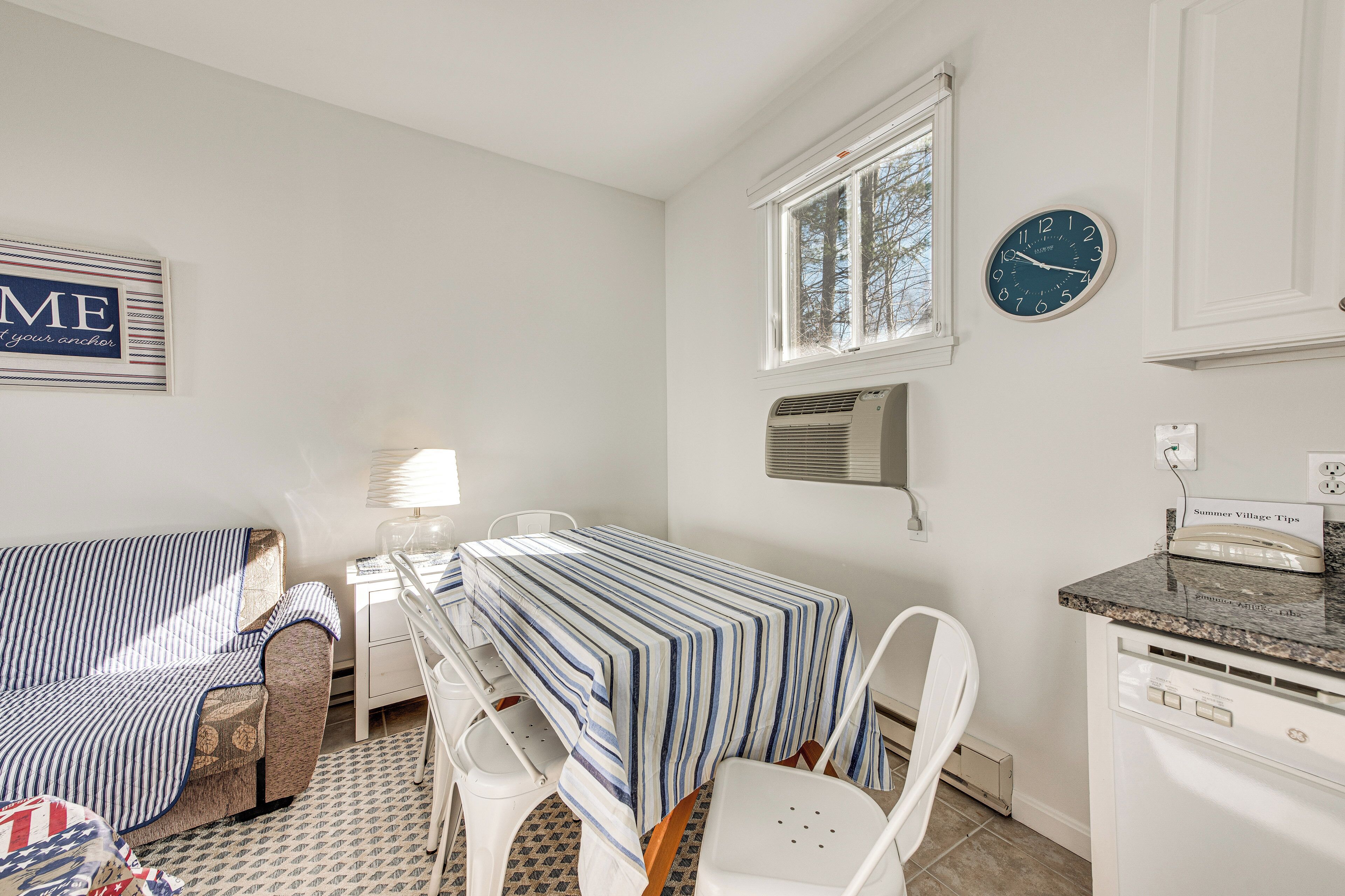 Una habitación con una mesa a rayas, sillas blancas, un sofá, un reloj y una ventana.