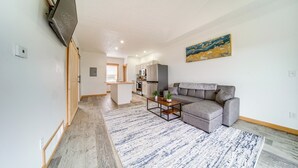 A modern living room with a grey sofa, a coffee table, and a blue and white rug. There is a kitchen in the background with white cabinets and a grey refrigerator. - Balcony+ Golf Course View: Quiet Erie Getaway (Erie)