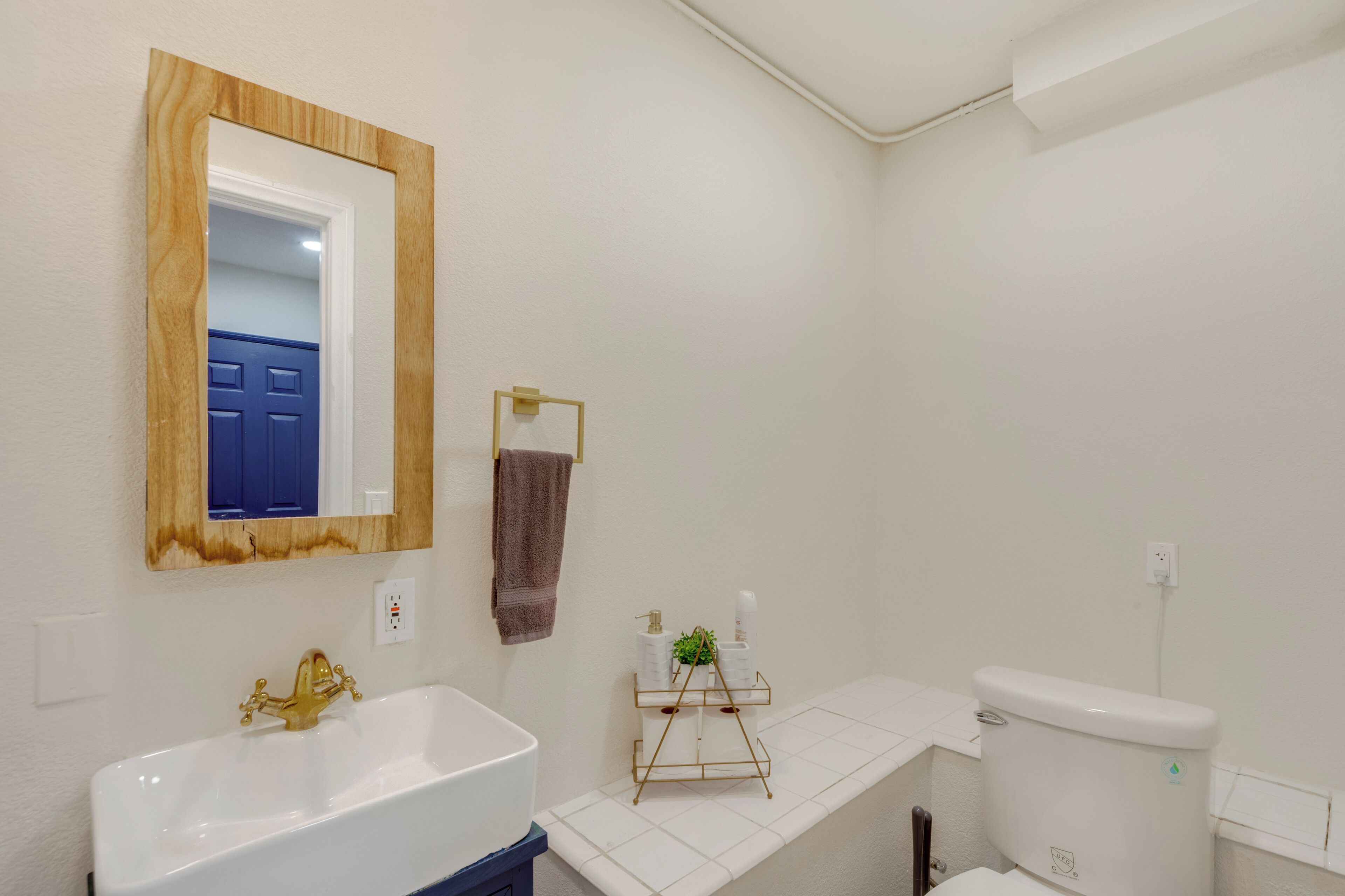 Ein Badezimmer mit einem weißen Waschbecken, einem goldenen Wasserhahn, einem Spiegel mit Holzrahmen, einer blauen Tür und einer weißen Toilette.