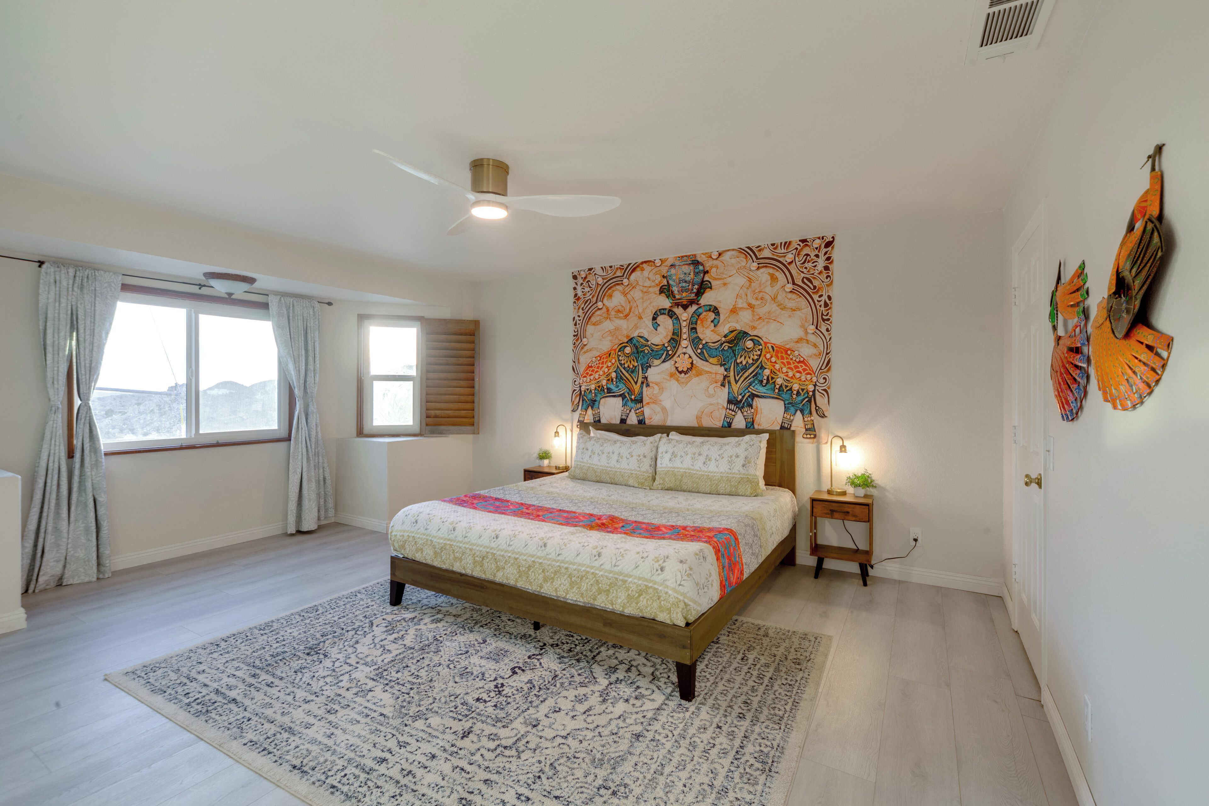 A bedroom with a large bed, a ceiling fan, a nightstand with a lamp, and a window with curtains.