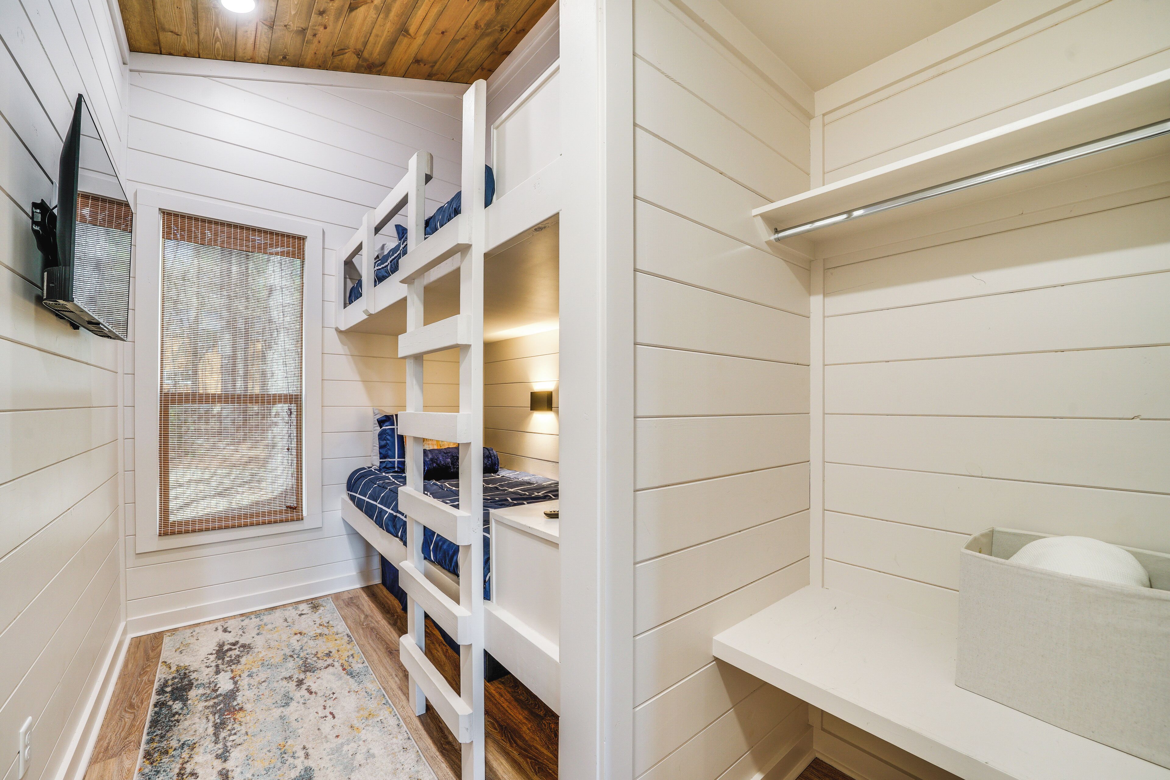 A compact room with bunk beds, a small window with blinds, a wooden ceiling, and a white wall.