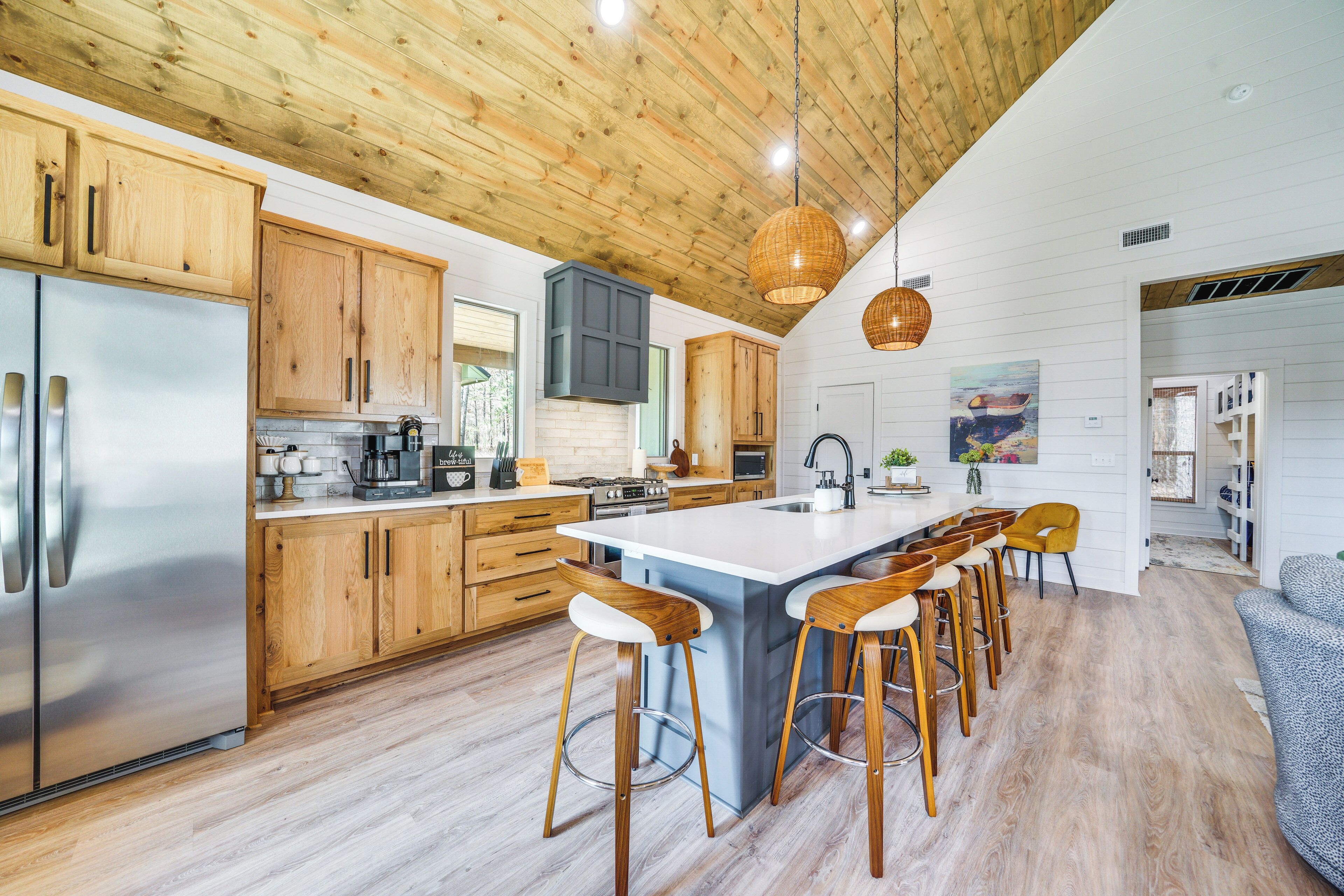 A modern kitchen with wooden cabinets, a stainless steel refrigerator, a large island with bar stools, and a hanging light fixture.