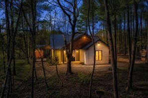 Une cabane aux murs en pierre, dotée d’une terrasse en bois et d’un toit en métal, entourée d’arbres.