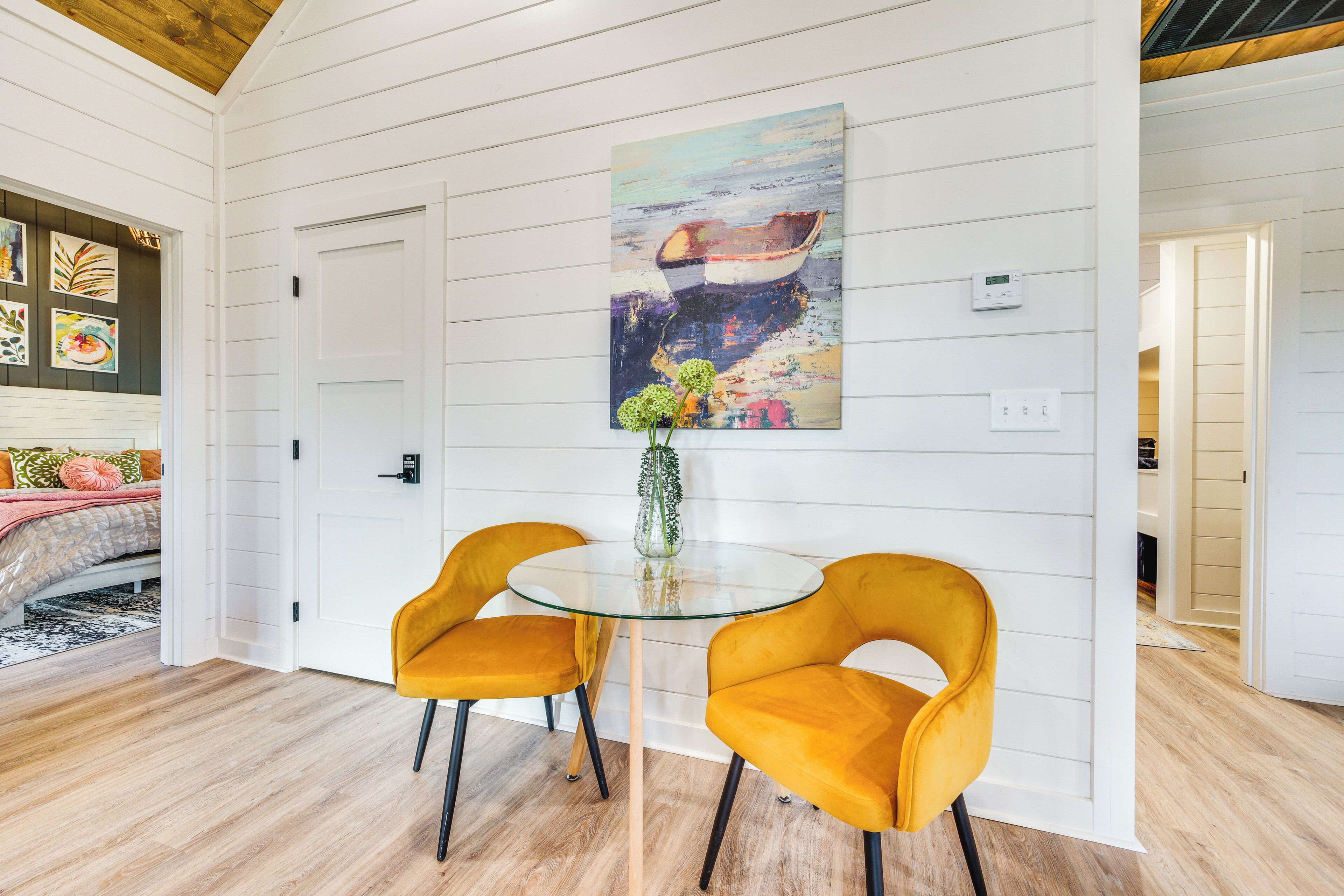 A dining area with a glass table and two yellow chairs, a painting on the wall, and a white door.
