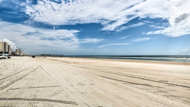 Ein weiter Sandstrand mit deutlich sichtbaren Spuren von Autoreifen, ein Steg in der Ferne und Gebäude entlang der Küste.