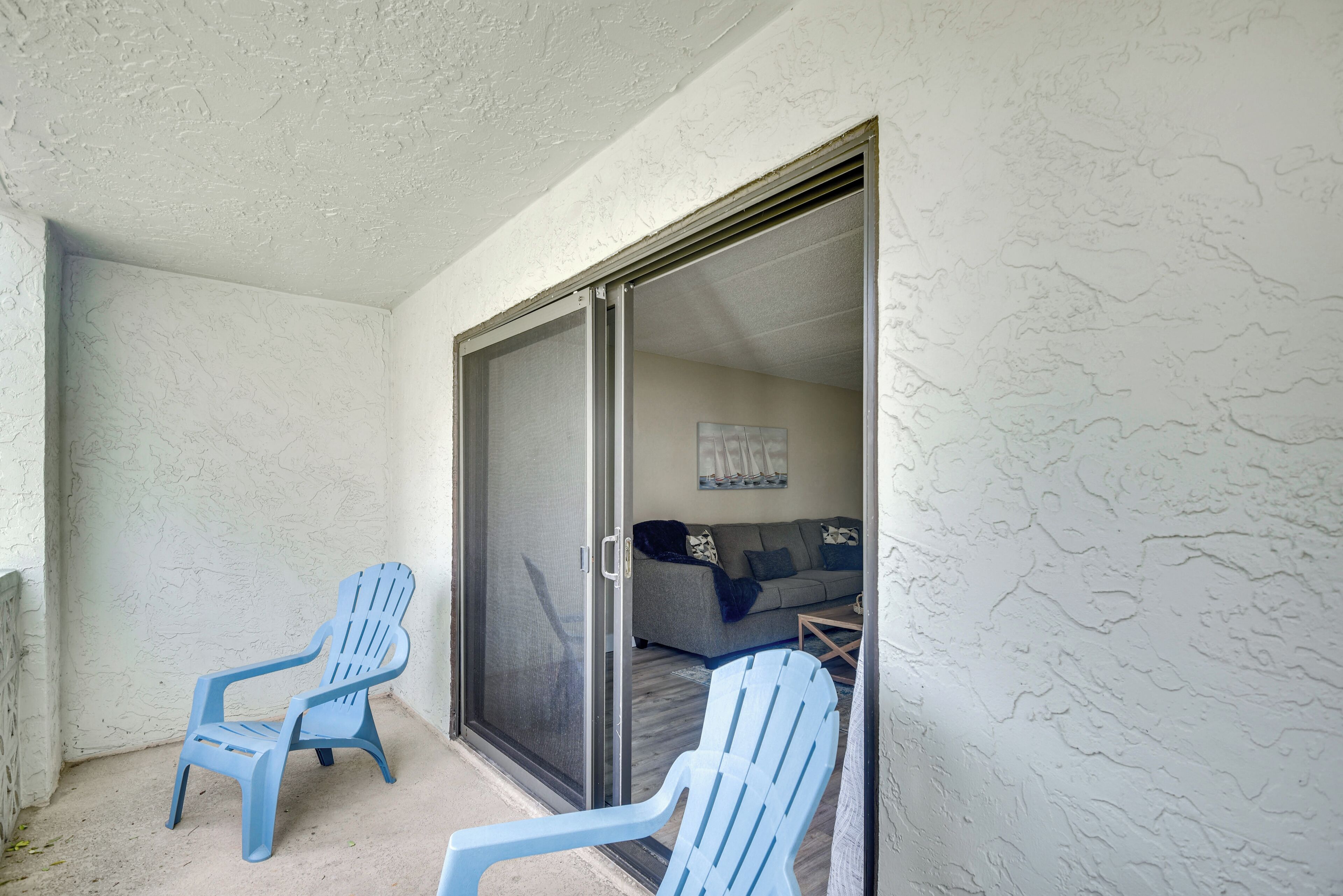 A balcony with two blue chairs, a sliding glass door leading to a living area with a sofa and a painting.