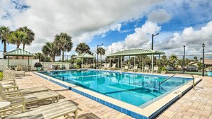 Ein rechteckiger Pool mit Fliesenrand, umgeben von Liegestühlen und Sonnenschirmen, mit Palmen und blauem Himmel im Hintergrund.