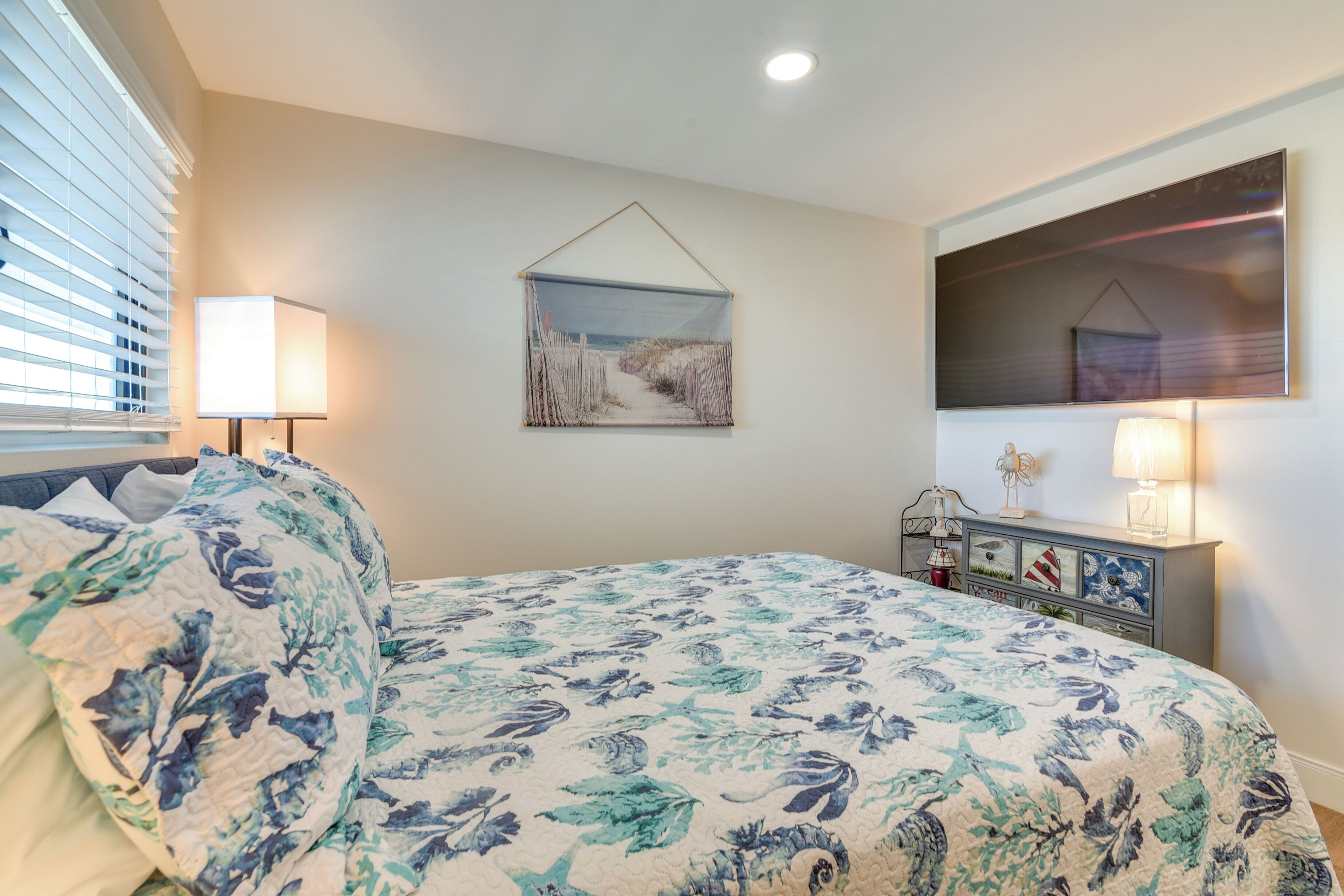 A bedroom with a floral bedspread, a bedside lamp, a dresser with decorative items, and a framed picture on the wall.