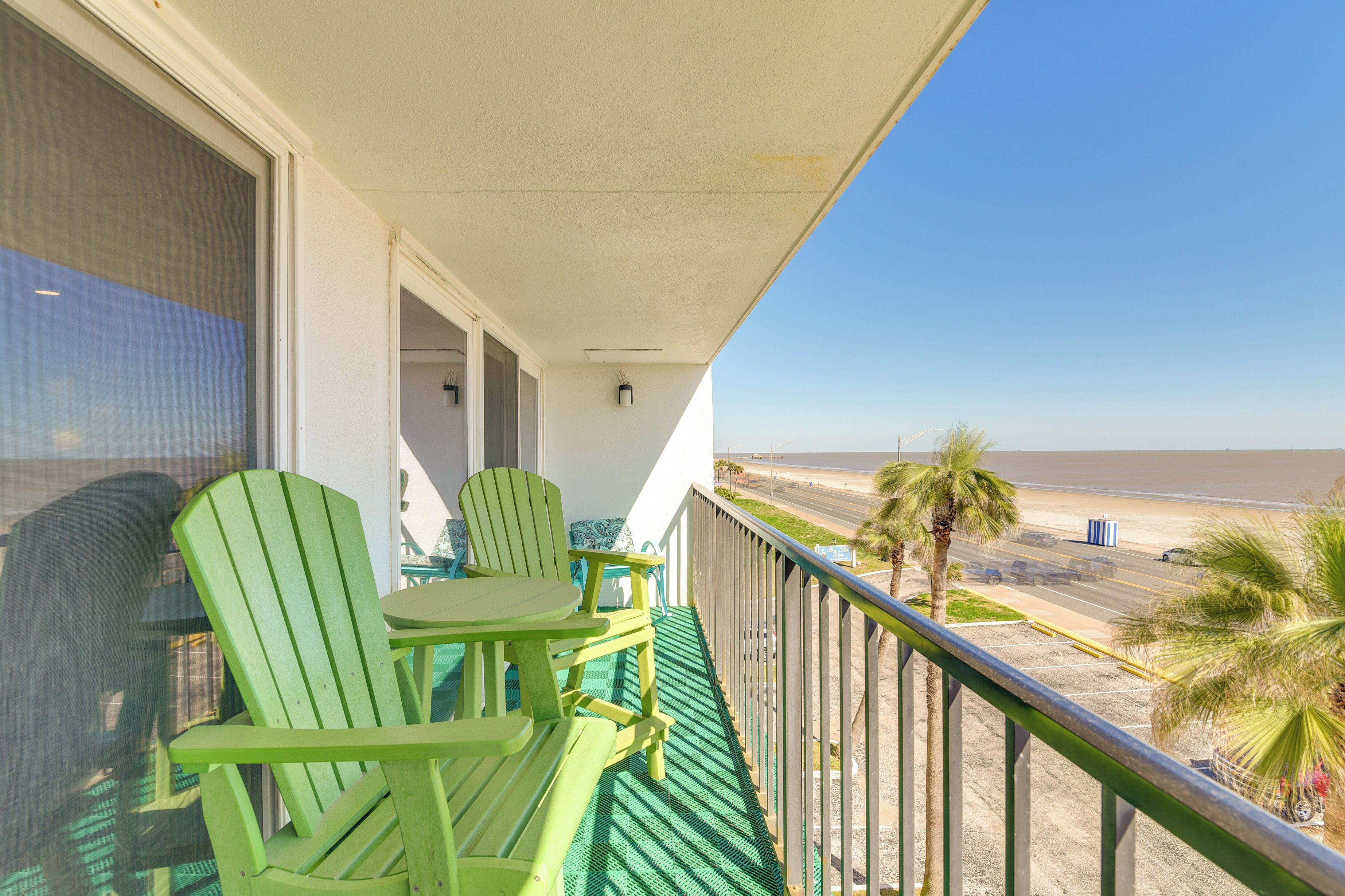 A balcony with green chairs and a table, overlooking a beach with palm trees and a road.