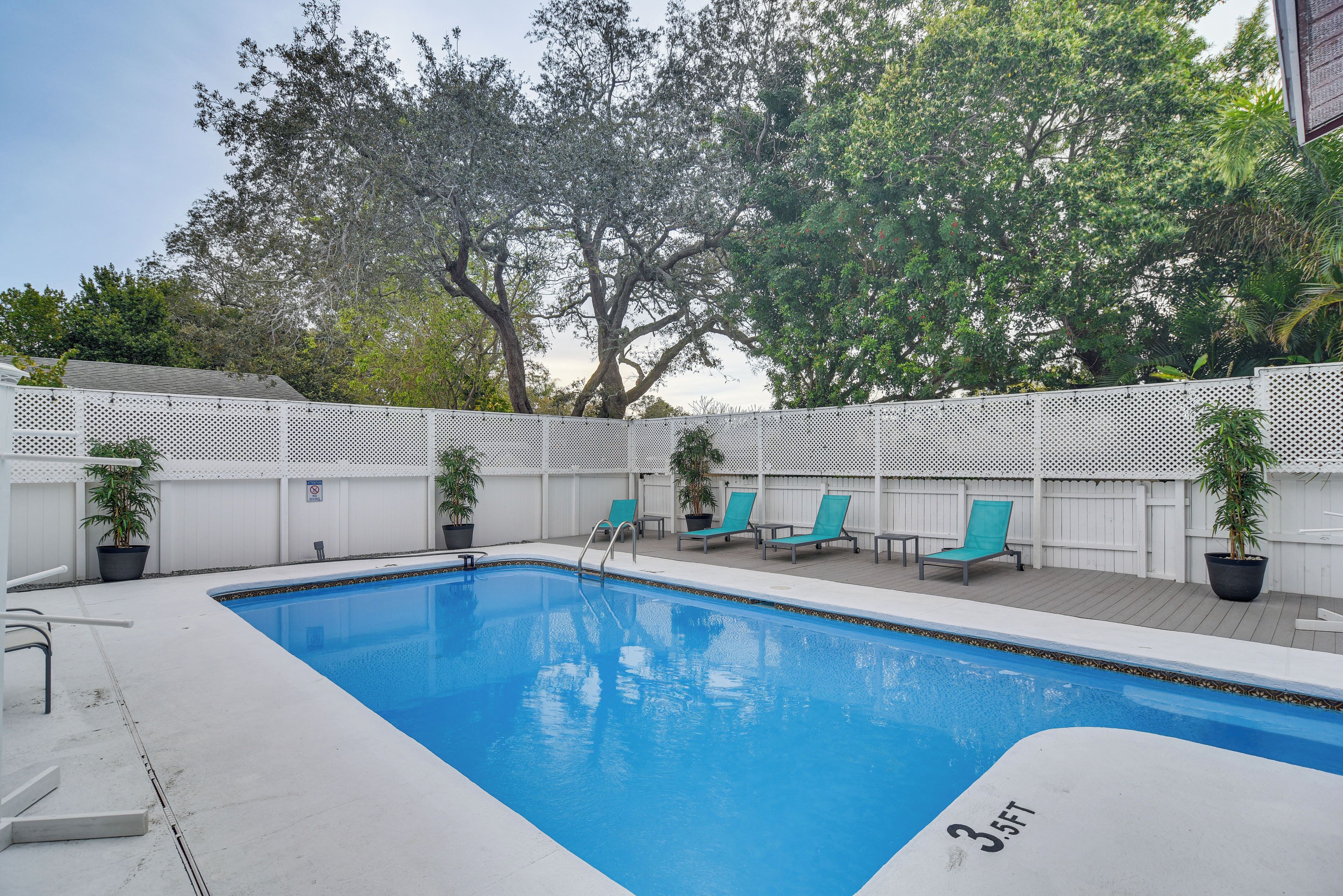 A rectangular swimming pool with a white edge, surrounded by a white fence, and three green lounge chairs.