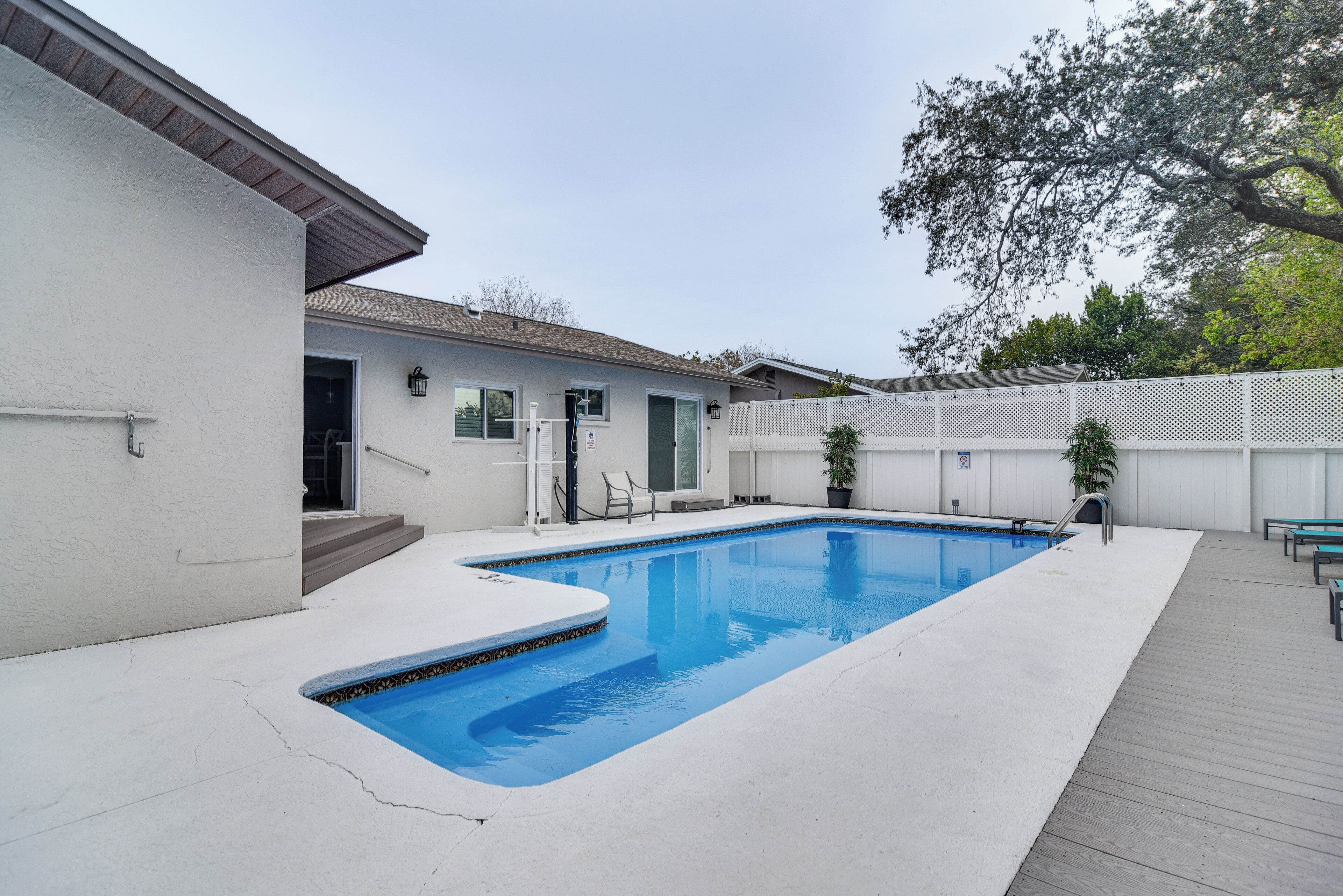 A rectangular swimming pool with a white deck, a covered patio area, and a white fence surrounding the property.