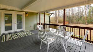 Une terrasse extérieure couverte, dotée d’une table à plateau de verre et de chaises blanches, d’une rambarde en bois et offrant une vue sur les arbres.