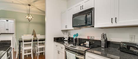 A modern kitchen with white cabinetry, a black countertop, stainless steel appliances, and a dining area with white chairs and a table.