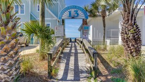 A wooden boardwalk leading to a beach access sign with palm trees and a building in the background. - Panama City Beach Home: Steps to the Ocean! (Panama City Beach)